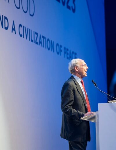 A man speaking at a Global Peace Convention podium in front of a large screen.